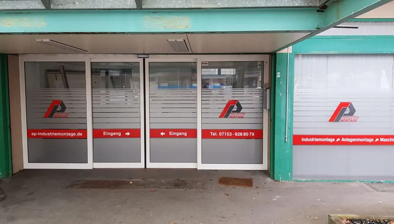 Automatische Glastüren eines Firmeneingangs mit Streifenfolie, Logo und Wegweiser zum Eingang
